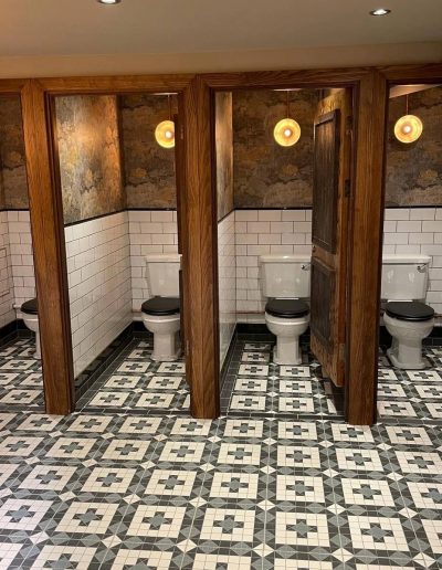 Four traditional black and white toilets with a green patterned tiled floor, illuinated by wall lights.